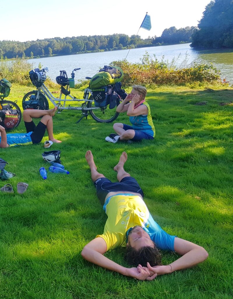 MT23_Entschleunigung_Natur_Pause Drei Menschen sitzen und liegen auf einer Wiese am Fluss zwischen den Tandems und machen Pause
