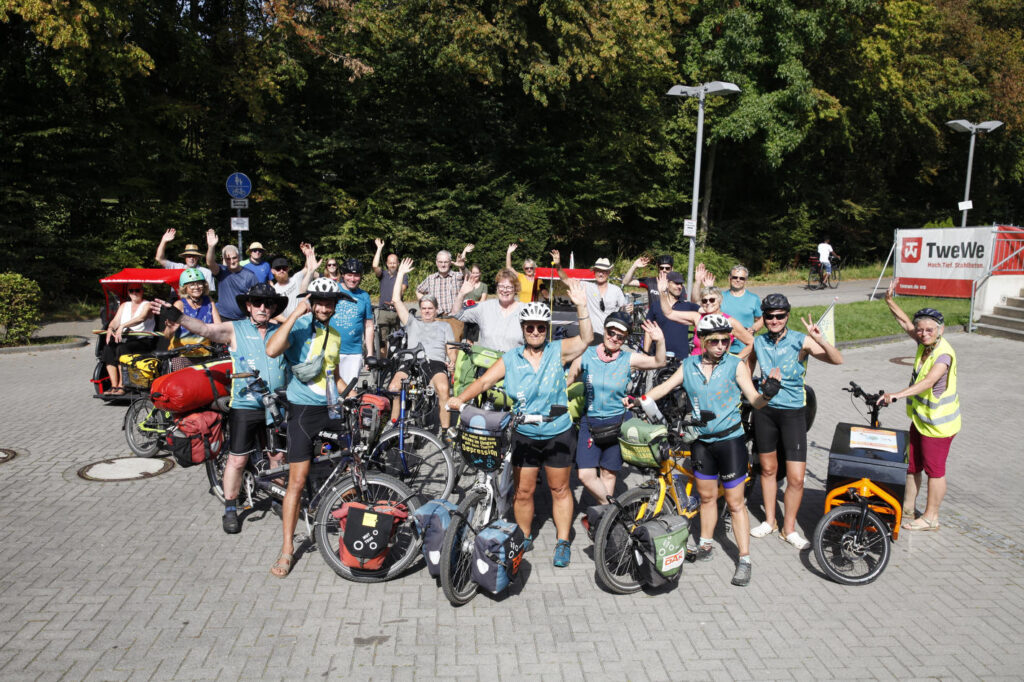 Eine Menschengruppe mit Tandems und MUT-TOUR-Trikots, die winken