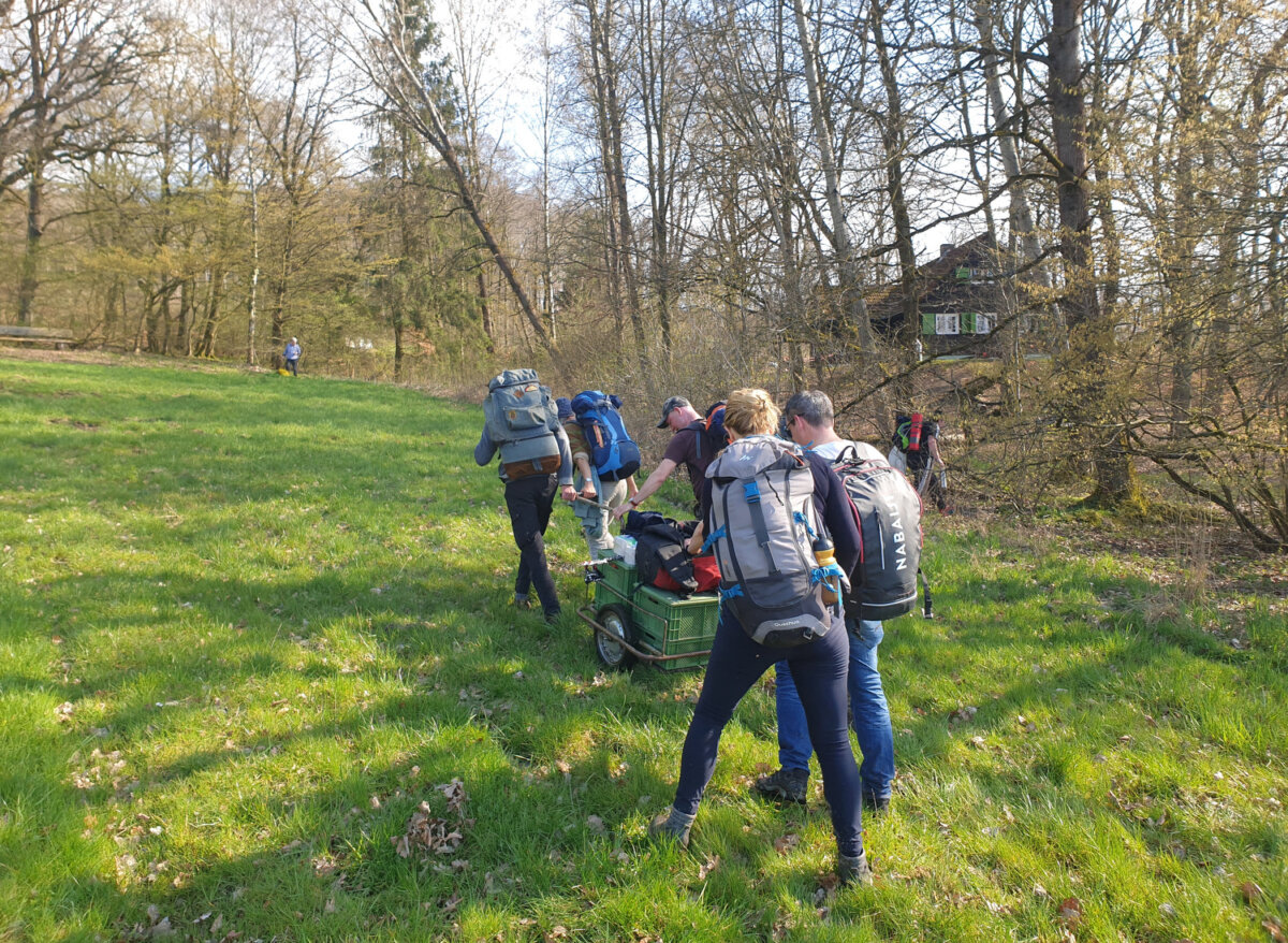 Fünf Menschen mit Rucksäcken ziehen einen Handwagen eine Wiese hinauf zu einem Haus
